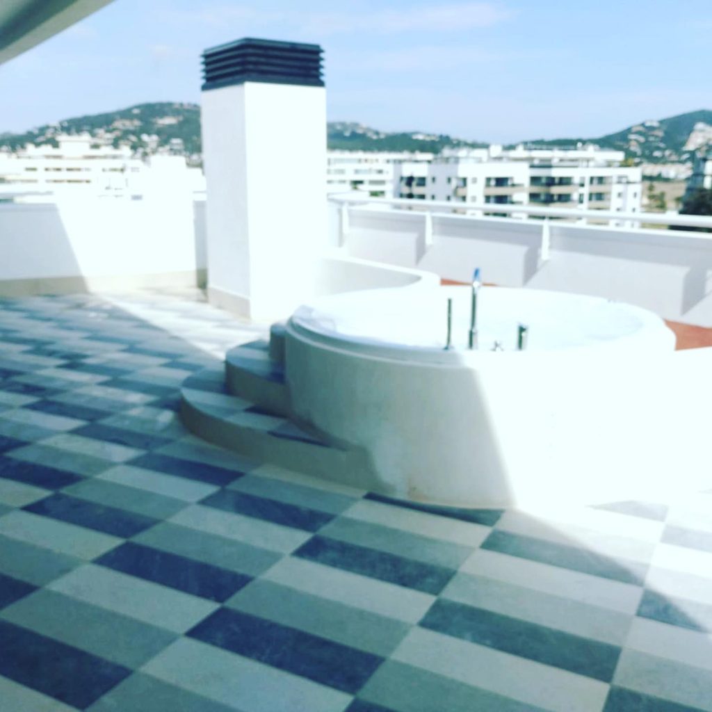 Terraza con diseño de baldosas modernas y un jacuzzi integrado, con vistas a edificios residenciales y montañas en Ibiza.